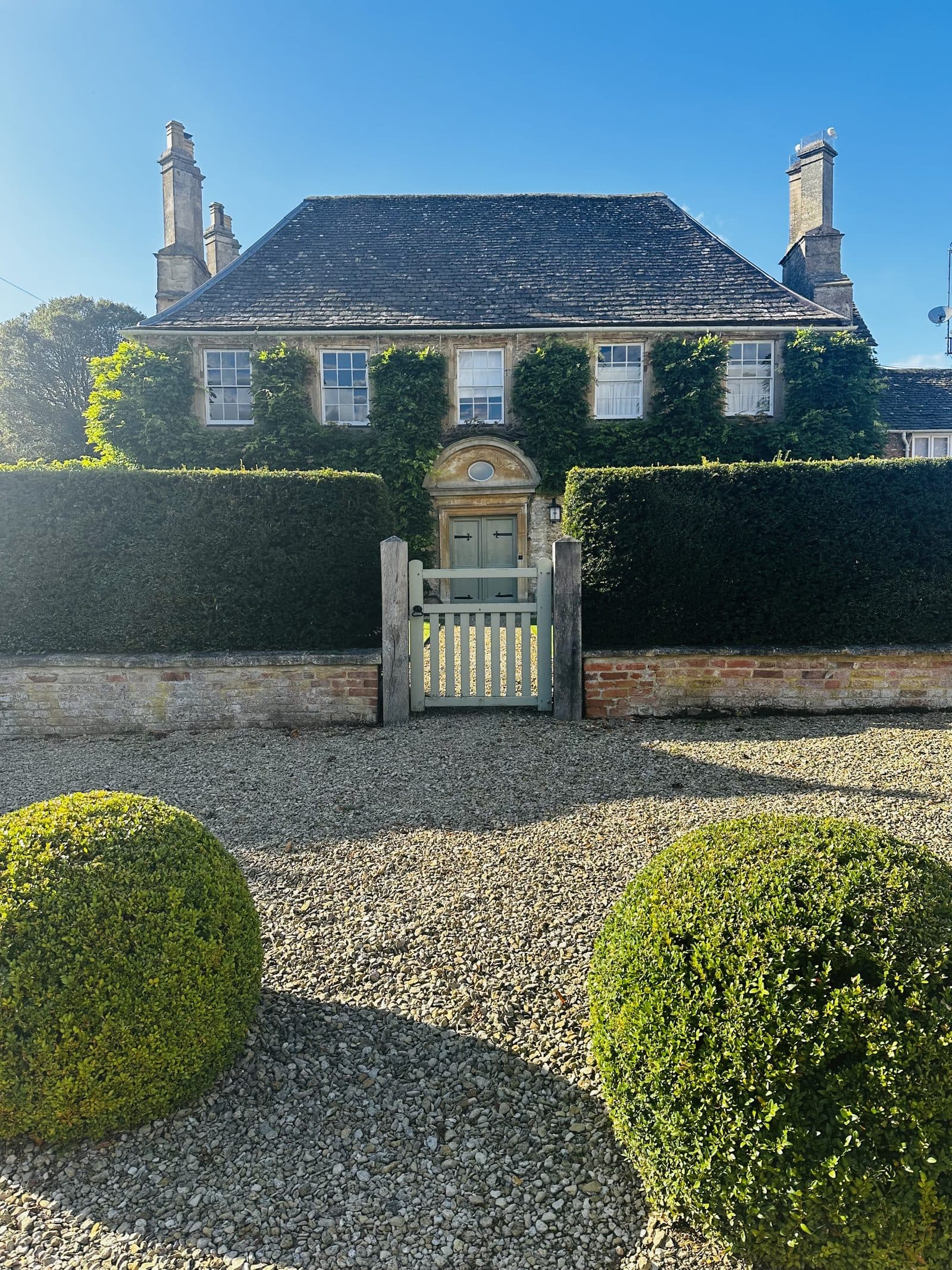 A clipped topiary hedge and gravel drive in front of a Cotswold stone house