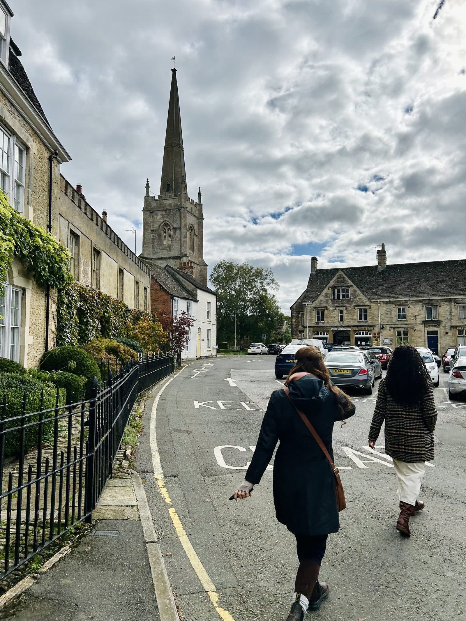 A Cotswolds village street curving downhill past stone houses under a cloudy sky