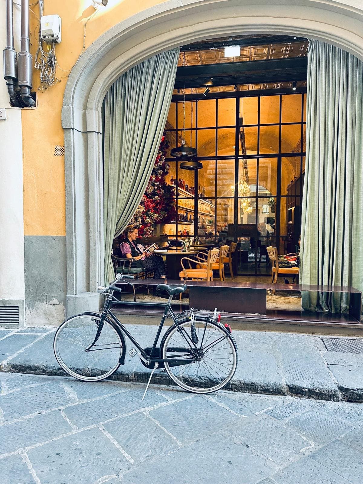 A bicycle propped against the kerb outside an arched Florence cafe with green curtains