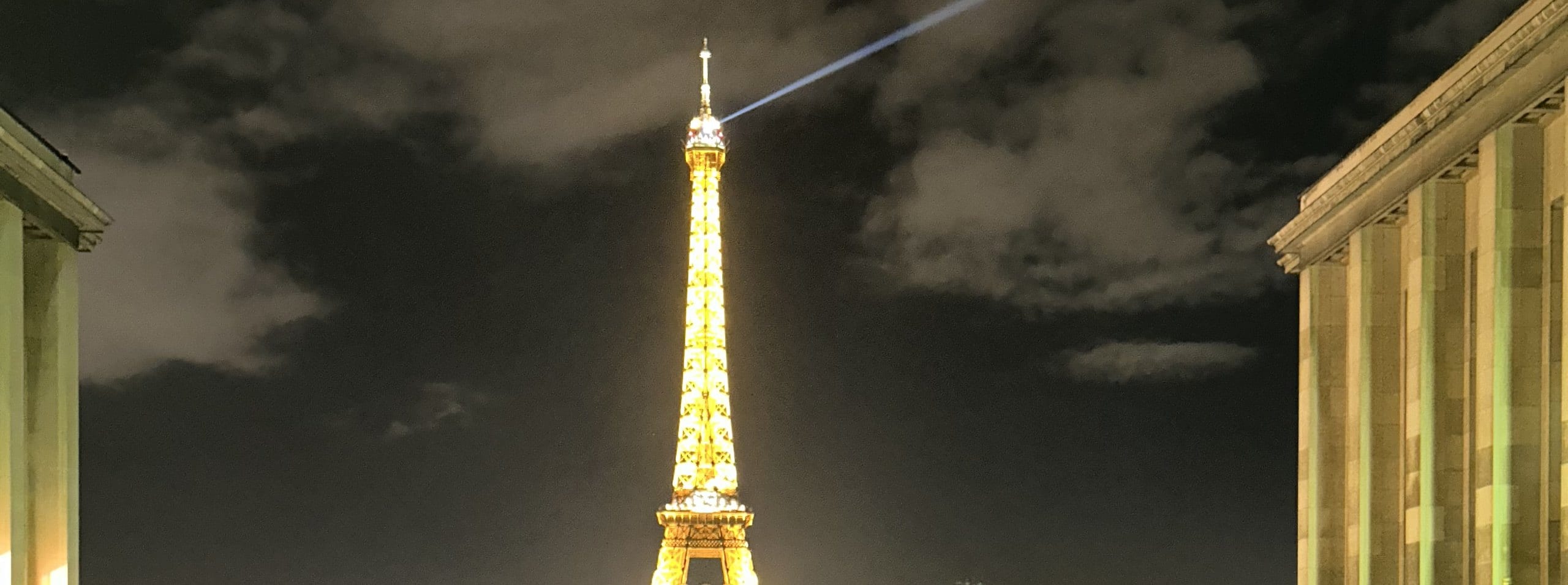 The Eiffel Tower lit gold at night, its beacon sweeping the clouds, framed by the colonnades of the Trocadéro