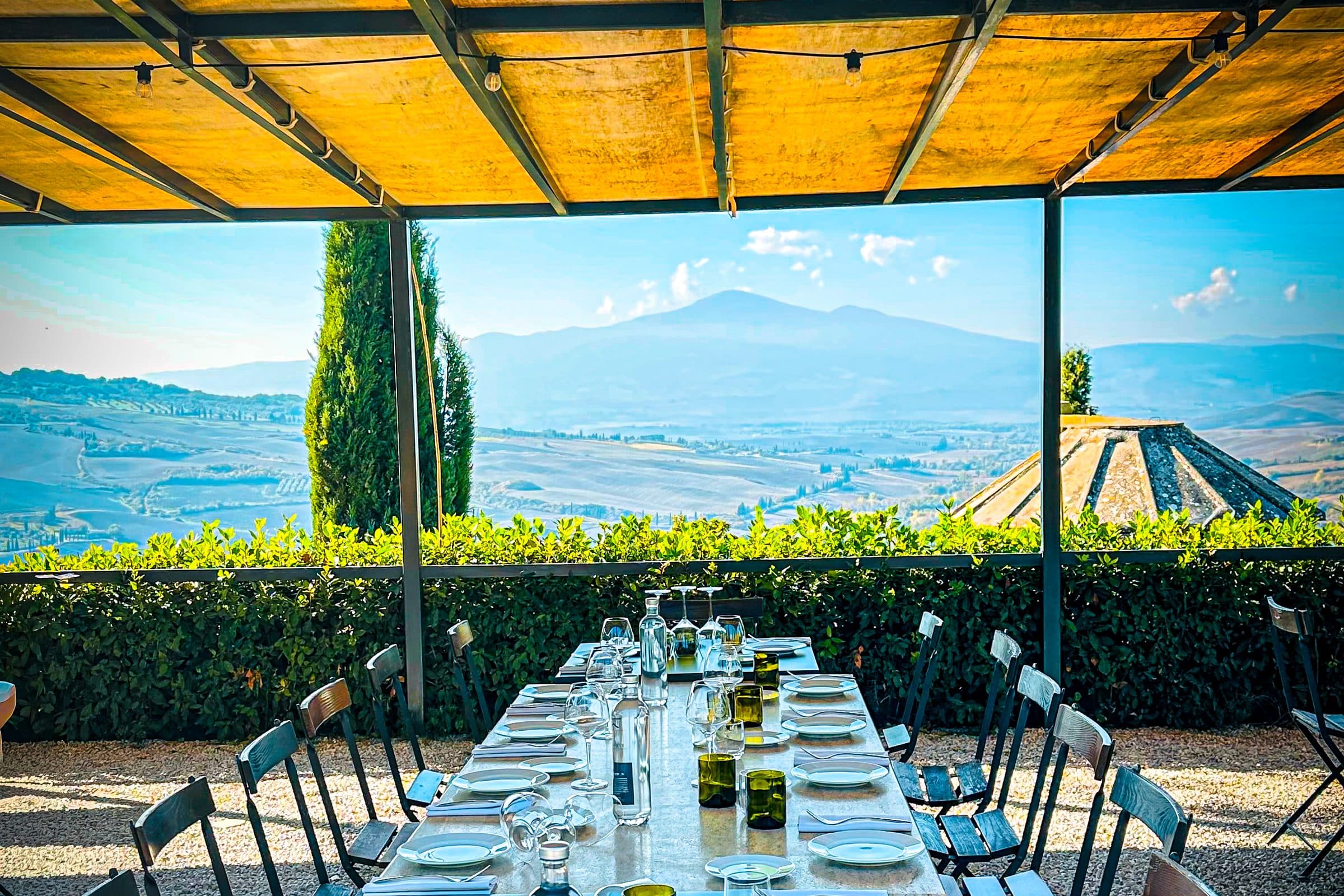A long table laid for lunch under a pergola, looking out over cypress trees and the Tuscan hills
