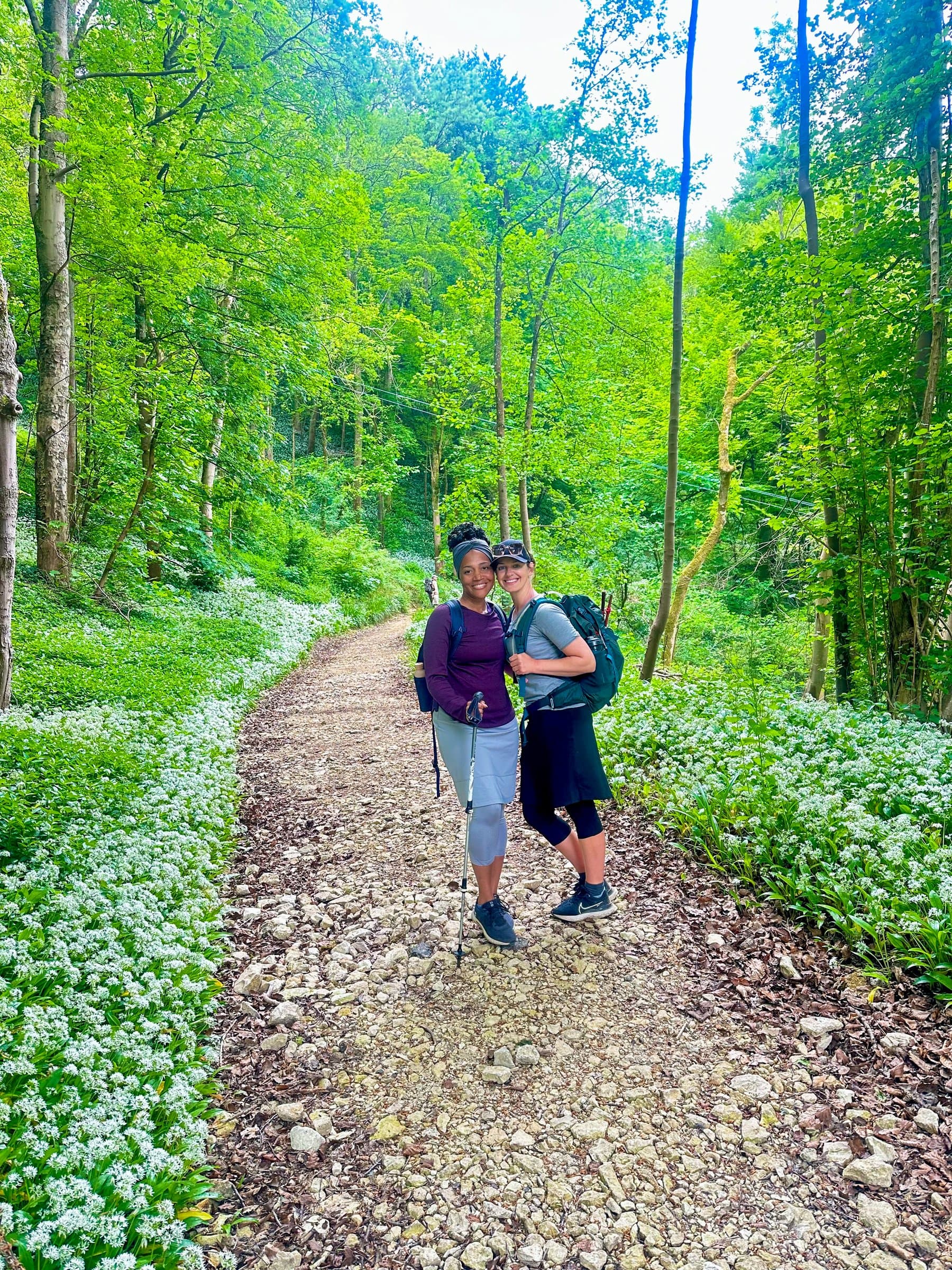 Two walkers with backpacks on a wooded trail carpeted with wild garlic