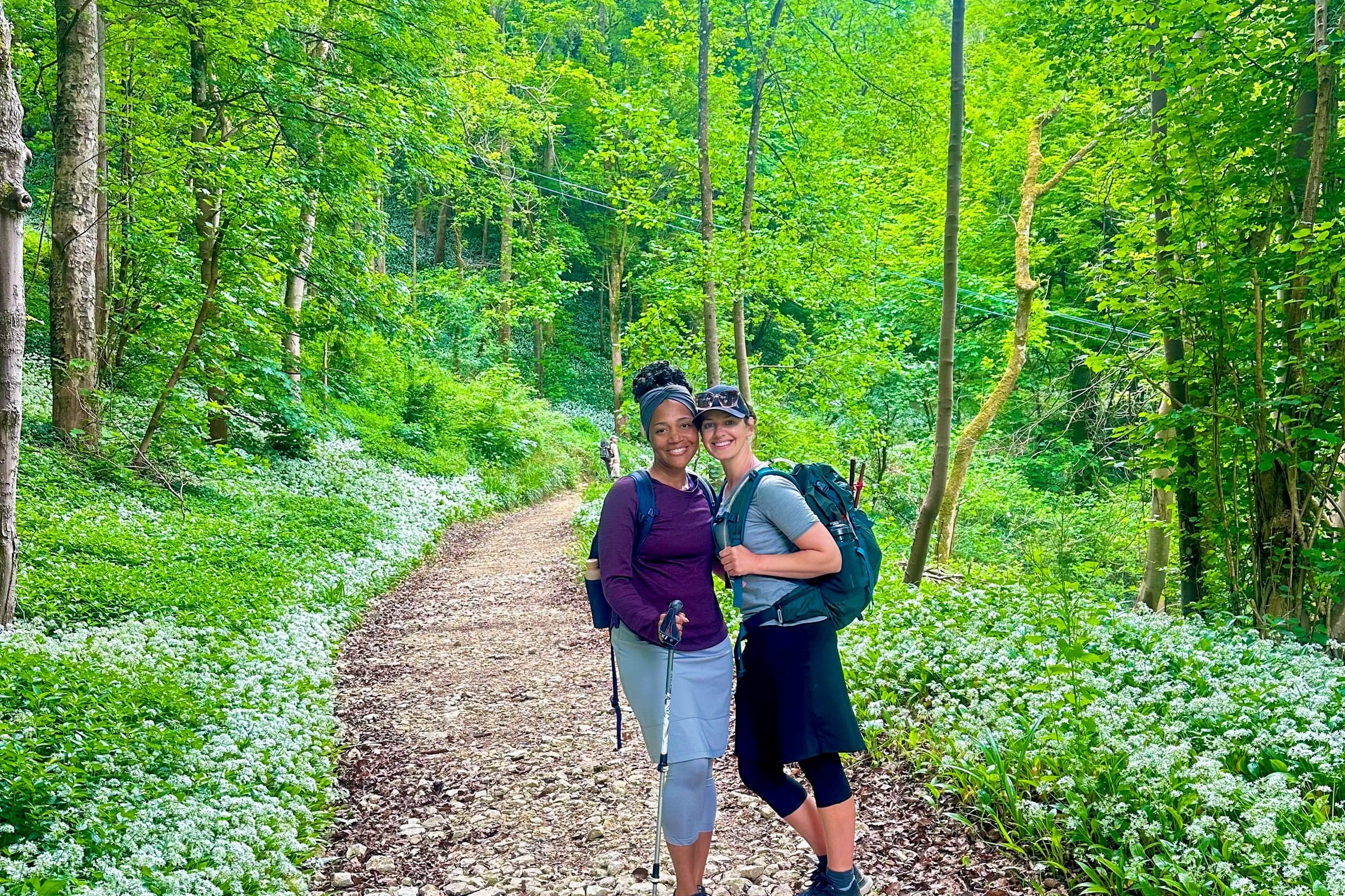 Two walkers with backpacks on a wooded trail carpeted with wild garlic