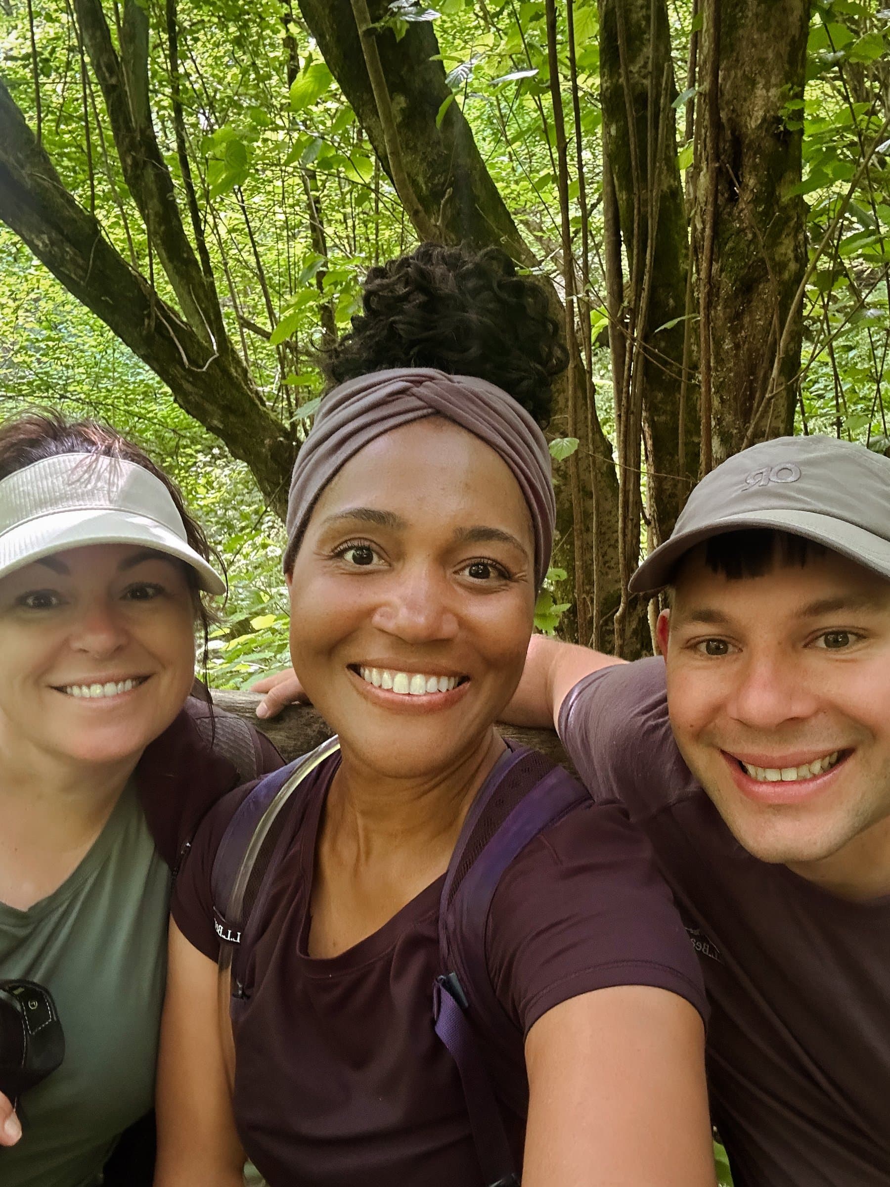 Three walkers with packs and caps pausing on a wooded trail