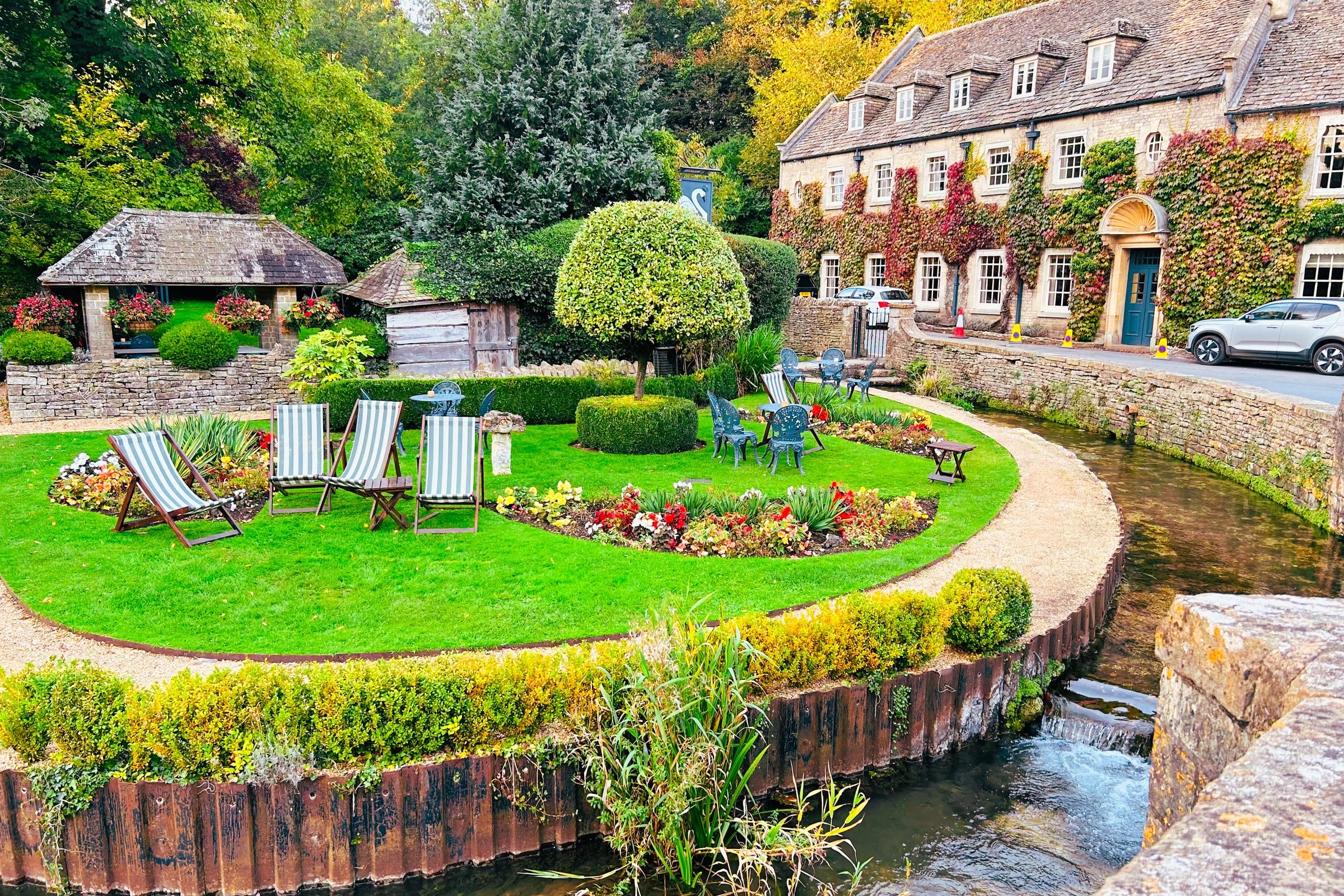 Deckchairs on a clipped lawn beside the stream at Bibury, with ivy-covered Cotswold stone cottages behind