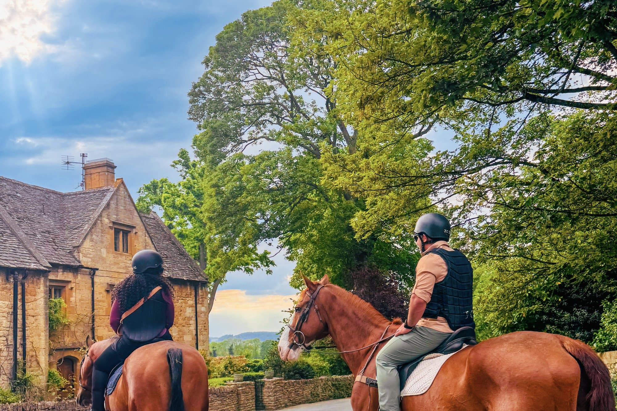 Two riders on horseback passing honey-colored stone cottages on a quiet Cotswolds lane