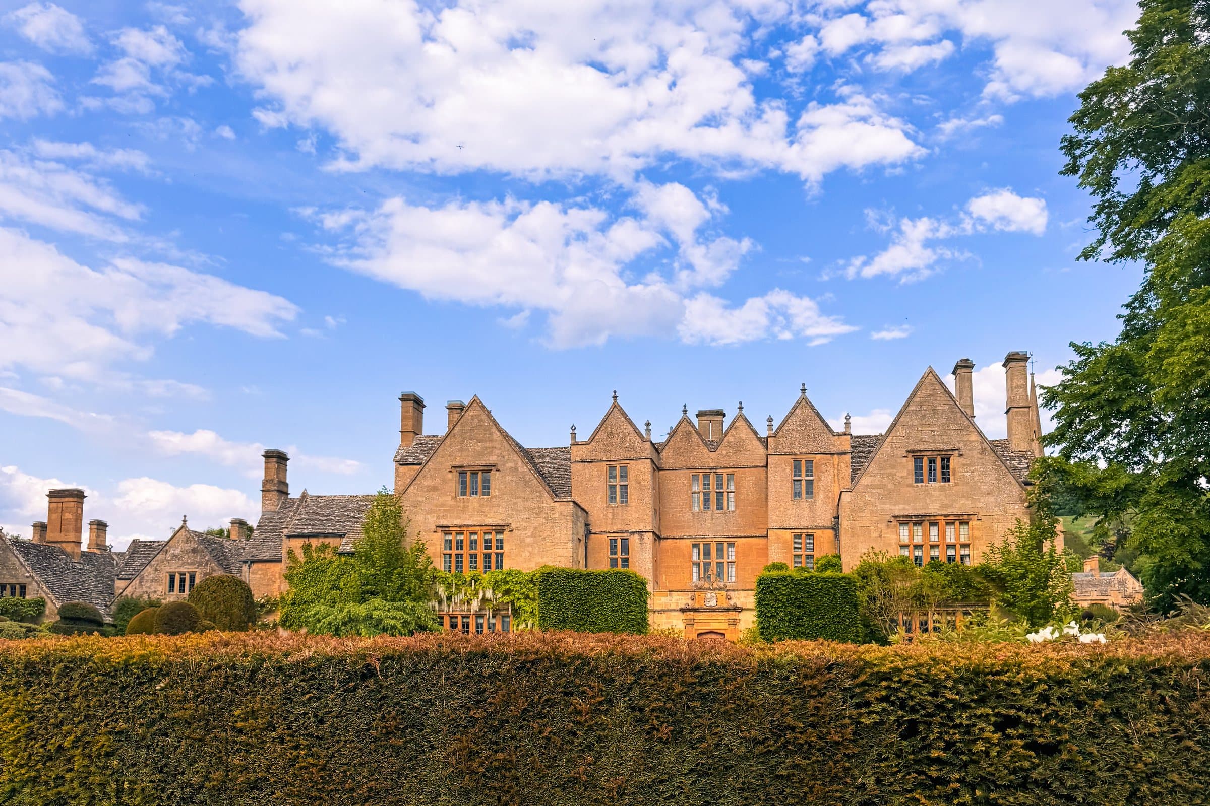 A gabled Cotswolds manor house behind a tall clipped hedge under a bright sky