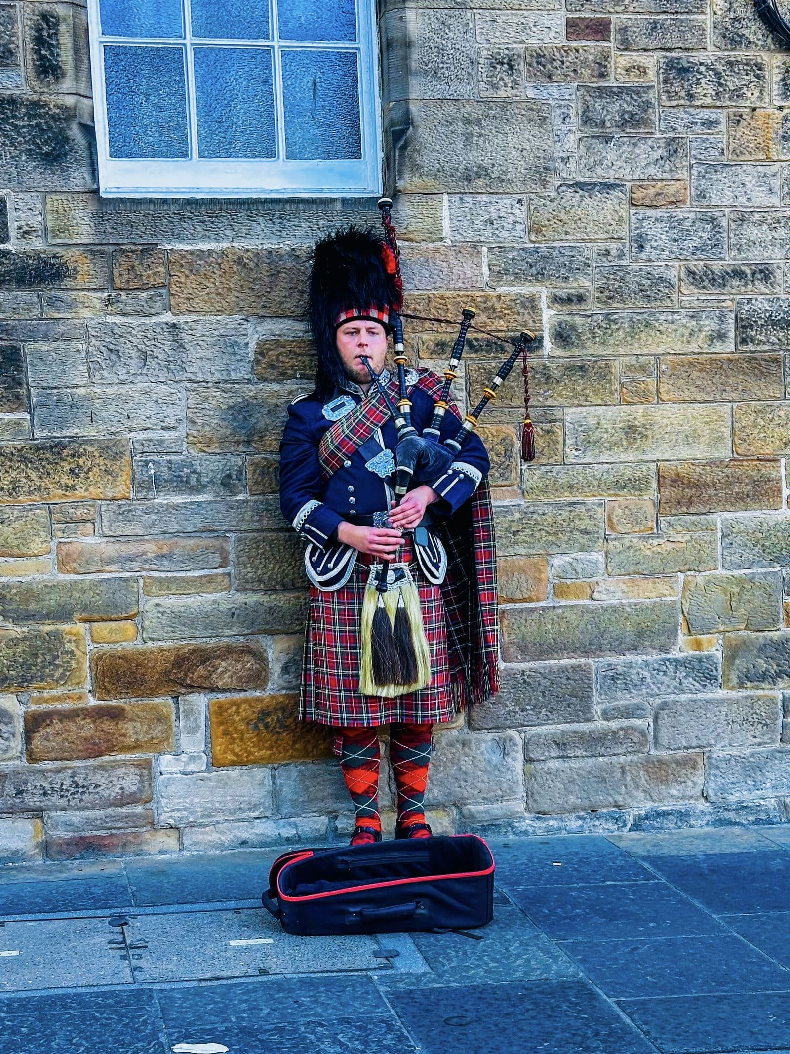 A piper in full Highland dress playing beside a stone wall in Edinburgh