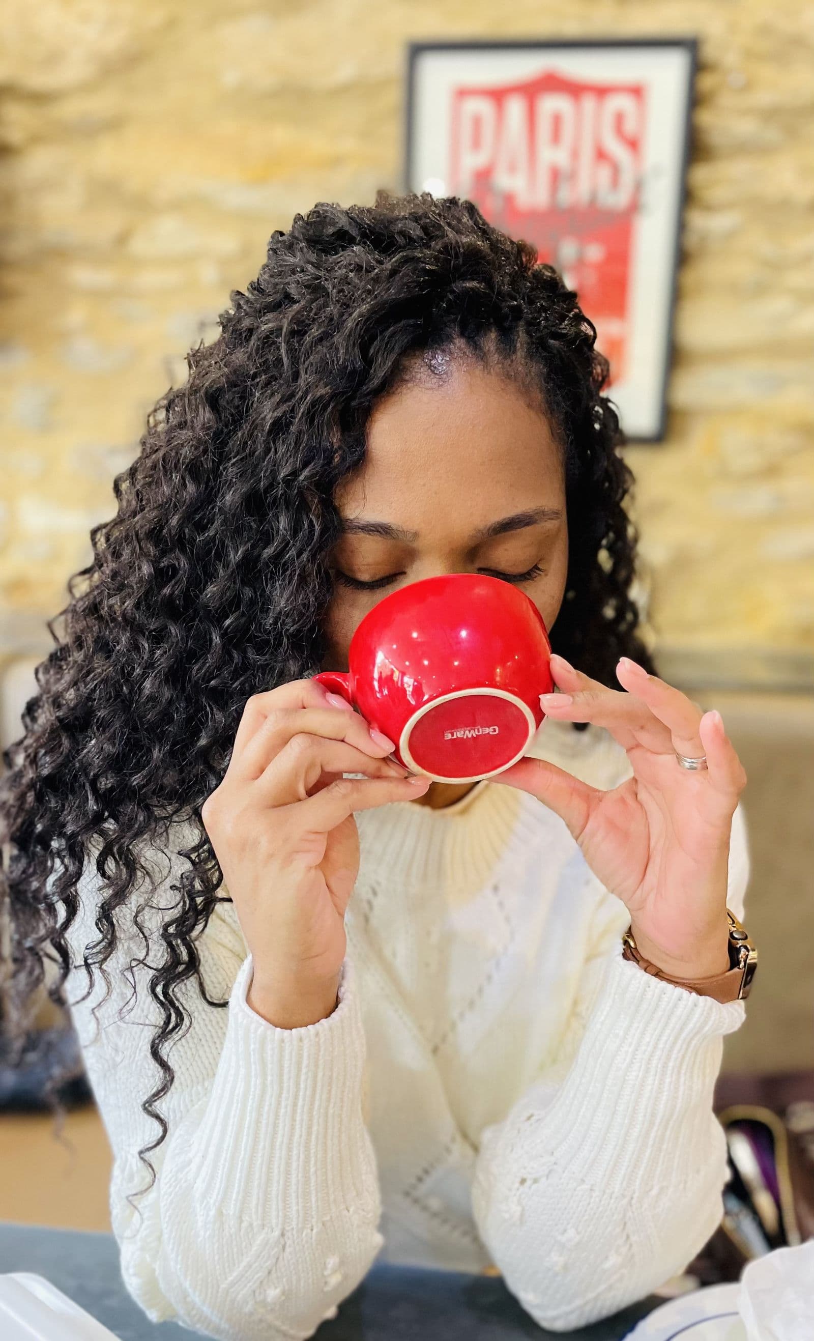 A red cup held in both hands at a cafe table beneath a Paris print