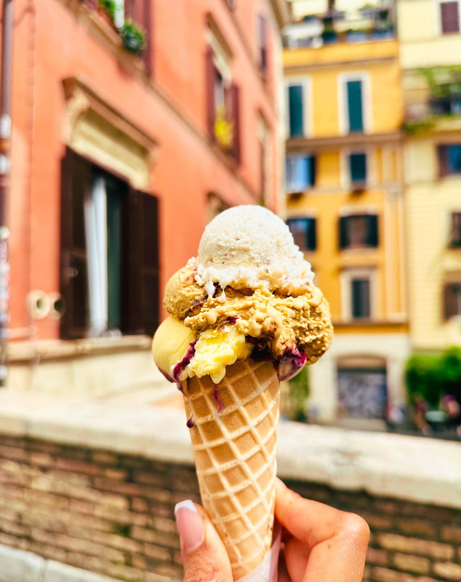 A gelato cone held up in front of a row of pink and orange Roman buildings