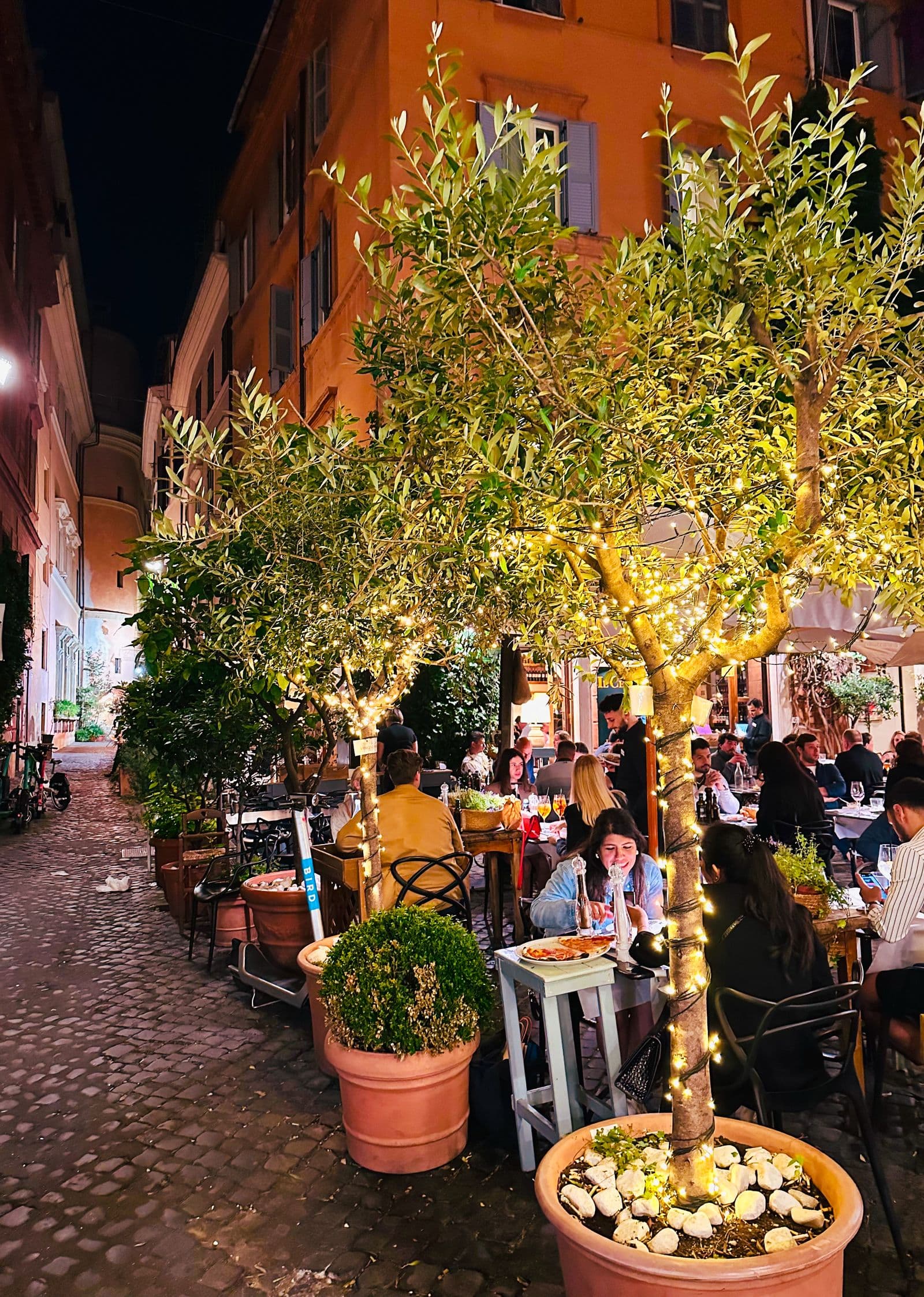 An evening street in Rome set with restaurant tables under strings of lights and potted olive trees