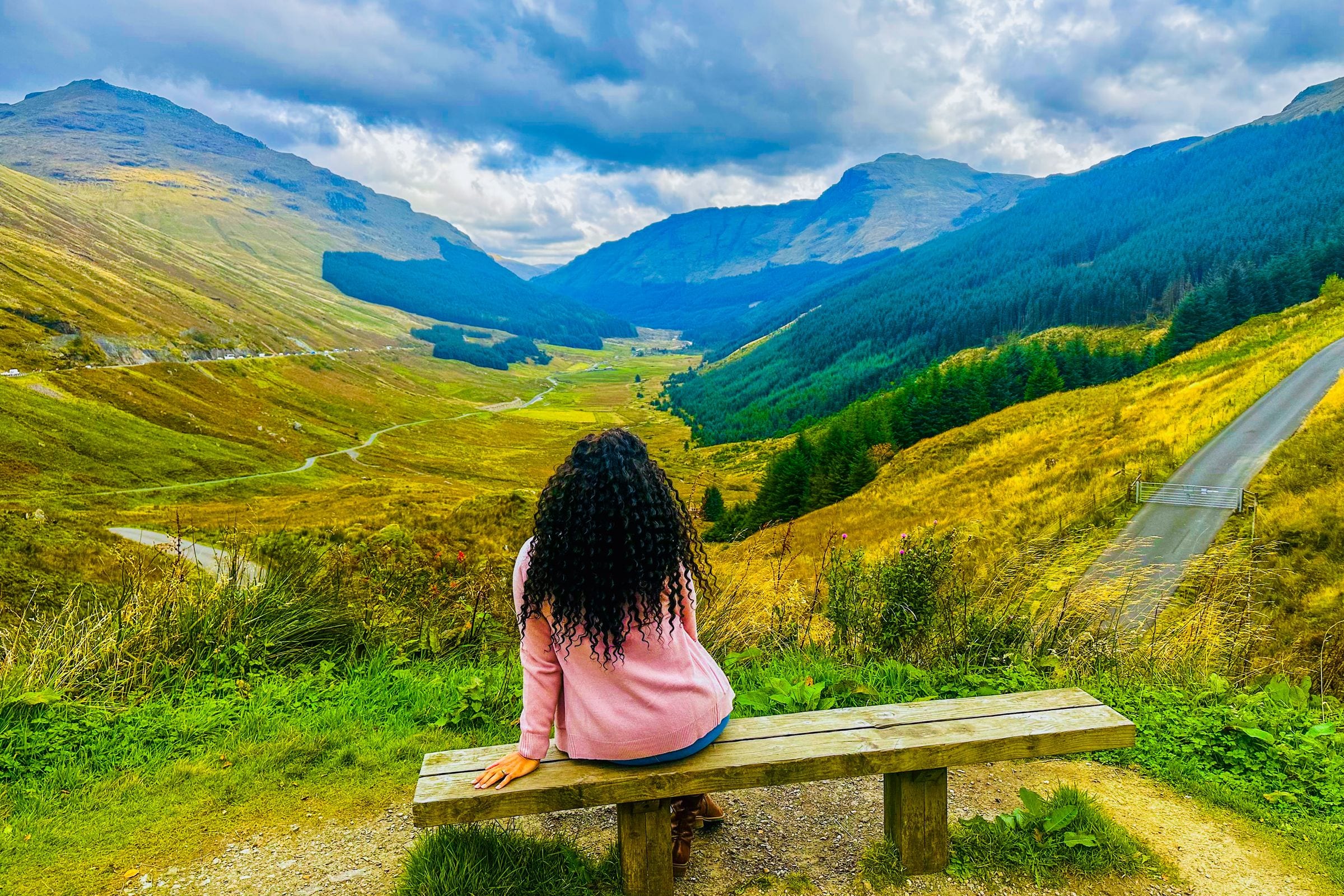 A traveler sitting alone on a wooden bench above a Highland glen, the road winding away between green and gold hillsides